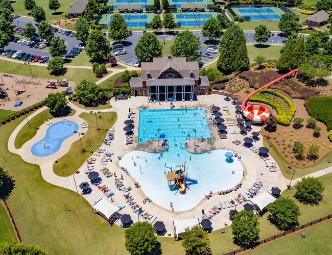 Aerial view of a vibrant community pool area with water slides, playgrounds, tennis courts, and surrounding greenery.